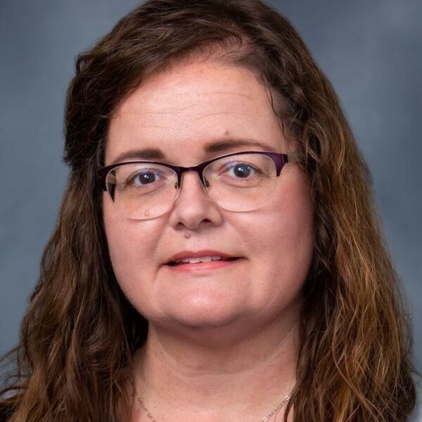 A smiling woman with long brown hair, purple glasses, and a silver necklace against a gray background.