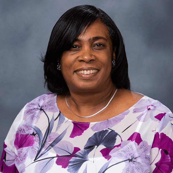 A smiling woman with dark skin and short dark hair wears a white shirt with purple and lavender floral patterns.