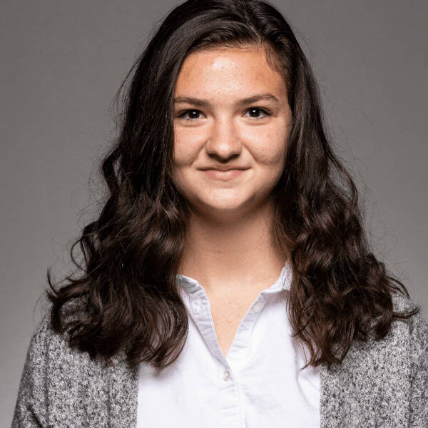Young woman with dark wavy hair, wearing a white collared shirt and gray speckled cardigan, smiling slightly.