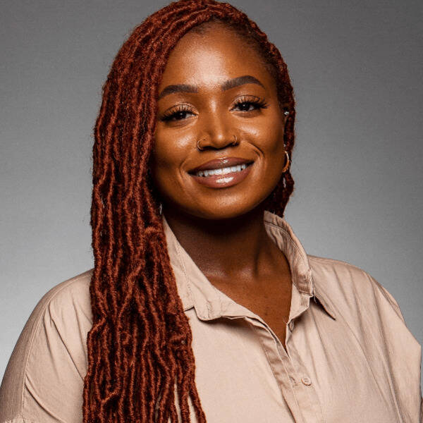 A woman with dark skin and long reddish-brown dreadlocks smiles warmly. She wears a tan collared shirt and has nose piercings.