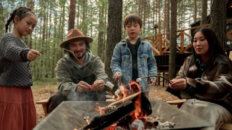 Four people, two adults and two children, roast marshmallows over a fire pit in a pine forest; the man smiles.
