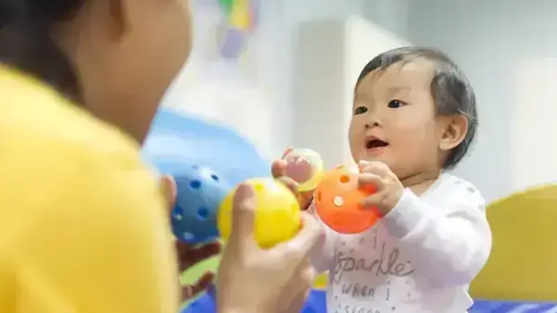 A smiling baby with dark hair holds an orange ball, looking at an adult in a yellow shirt holding blue and yellow balls.