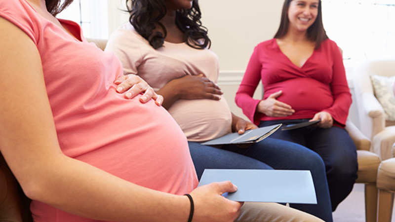 Three pregnant women smile while sitting in a group, gently touching their bellies and holding papers.