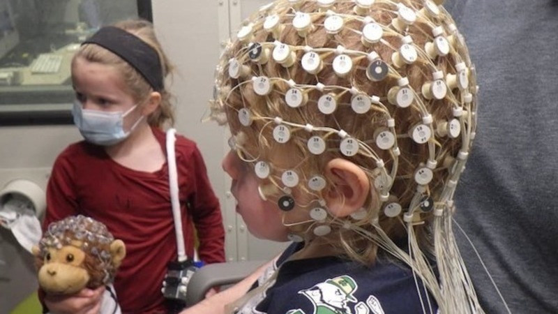 Child with EEG cap and Fighting Irish shirt observed by masked child holding a stuffed monkey.