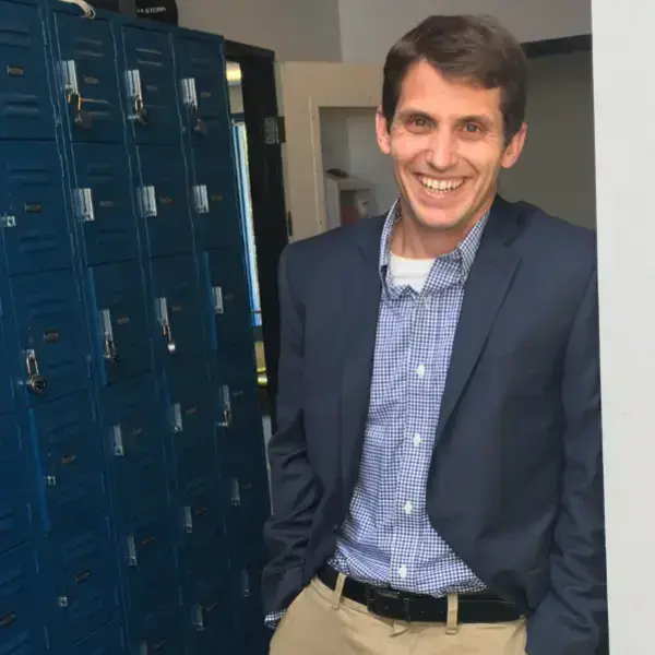 A smiling man in a navy blazer, light blue gingham shirt, and khaki pants stands with hands in pockets by blue lockers.