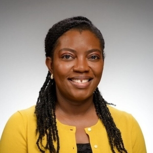 A smiling Black woman with braided hair, a gap in her front teeth, wearing a yellow cardigan and white earrings.